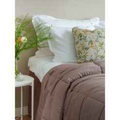 Kuvassa näkyy osa sängyn päädystä. Sängyssä on maitokahvin värinen topattu päiväpeitto, kukallinen koristetyyny ja valkoiset tyynyt. Sängyn vieressä yöpöydällä on kukkia ja vihreitä oksia maljakossa.